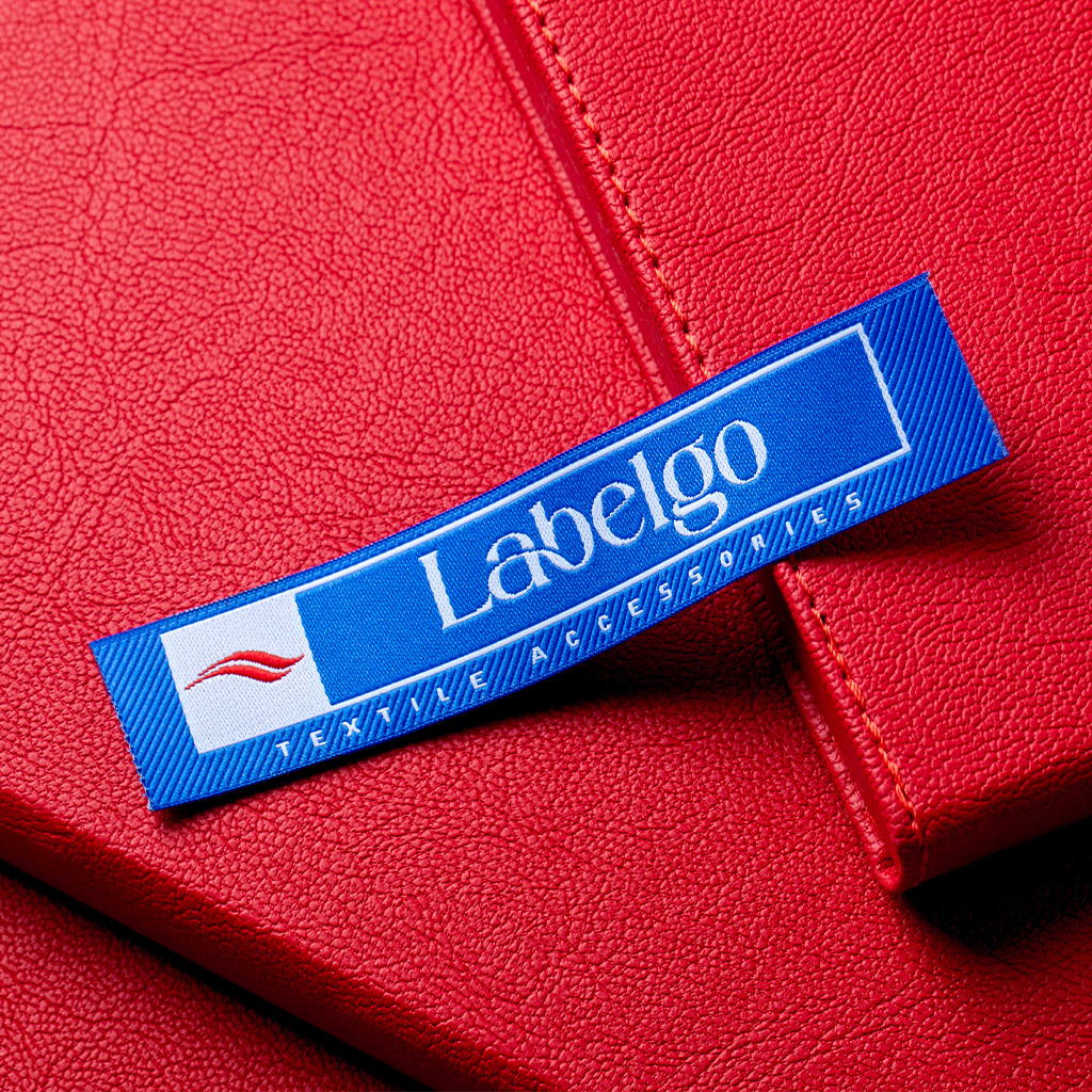 Etichetta tessuta blu accesa con logo bianco posizionata su una superficie in pelle rossa.